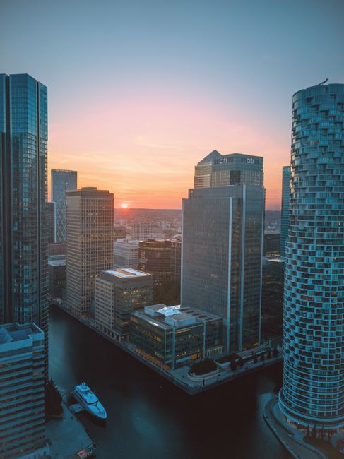 A high-angle view of a cityscape at sunset featuring modern skyscrapers with reflective glass facades surrounding a narrow waterway. In the foreground, a white boat is docked along the water's edge near the base of the buildings. The sky displays a gradient from soft pink to light blue, indicating the approaching evening. The scene captures the urban environment typical of Canary Wharf, with tall office buildings and residential towers. This image is relevant to home relocation and furniture transport services offered by Canary Wharf Man and Van, illustrating the bustling city environment where house removals and moving logistics take place, particularly as part of professional packing, loading, and transportation operations within the district.