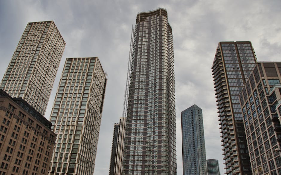 A high-angle view of a cityscape at sunset featuring modern skyscrapers with reflective glass facades surrounding a narrow waterway. In the foreground, a white boat is docked along the water's edge near the base of the buildings. The sky displays a gradient from soft pink to light blue, indicating the approaching evening. The scene captures the urban environment typical of Canary Wharf, with tall office buildings and residential towers. This image is relevant to home relocation and furniture transport services offered by Canary Wharf Man and Van, illustrating the bustling city environment where house removals and moving logistics take place, particularly as part of professional packing, loading, and transportation operations within the district.