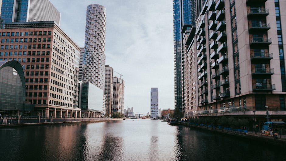 A wide view of Canary Wharf in London showing the city skyline with modern high-rise office buildings on both sides of the river. The buildings feature glass facades, steel structures, and contemporary architectural designs. In the foreground, the calm water of the river reflects the surrounding skyscrapers. To the right, a van with open doors is visible, with a few packed cardboard boxes, bubble wrap, and furniture blankets nearby, indicating a home relocation or professional removals process. The loading area is on a paved walkway adjacent to the building, where movers from Canary Wharf Man and Van are engaged in loading or unloading furniture and boxes onto the vehicle. The scene captures the urban environment with clear daylight, highlighting the logistics involved in furniture transport and packing during a same-day move in Canary Wharf, E14.