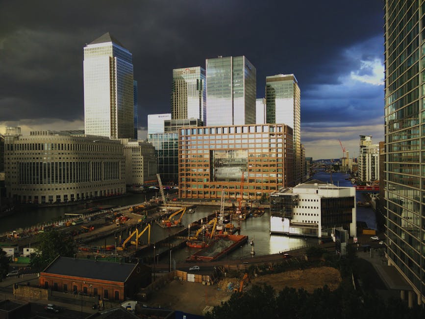 A cityscape view of Canary Wharf with modern high-rise office buildings, some with reflective glass facades, under a dark, partly cloudy sky. In the foreground, there is a construction site with multiple yellow cranes, construction vehicles, and materials such as wooden planks and metal scaffolding next to a waterway. Adjacent to the site, a small brick building is visible amongst the modern infrastructure. The scene is lit with natural daylight, highlighting the contrast between the ongoing construction and the sleek, finished buildings typically associated with professional house removals and furniture transport logistics. Canary Wharf Man and Van, a company specialising in removals, operates within this environment, supporting home relocation and moving services involving the careful handling and transportation of furniture and belongings.