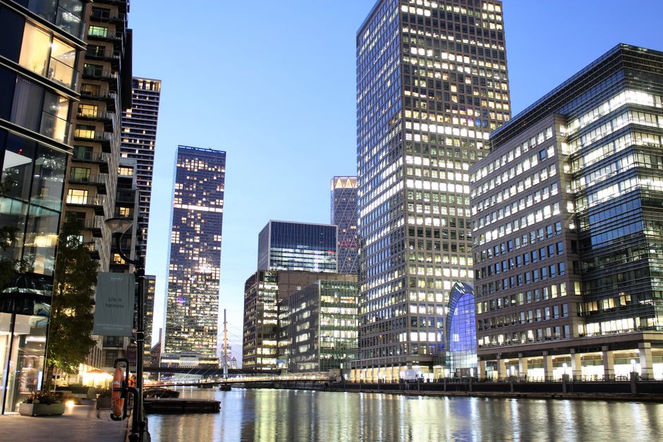 Photograph of a modern urban waterside scene during twilight, featuring a row of tall glass office buildings with illuminated windows reflecting the sky and surrounding structures. The buildings are positioned along a paved walkway by the water's edge, with some greenery and street lamps visible on the left side. The water in the foreground is calm, creating reflections of the buildings and lights. The scene is well-lit with natural and artificial lighting, and the overall environment suggests a city centre area in Canary Wharf, suitable for house removals and moving services such as furniture transport and packing during home relocations, as handled by Canary Wharf Man and Van.