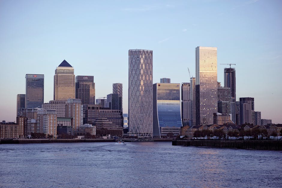 A cityscape view of Canary Wharf with modern high-rise office buildings, some with reflective glass facades, under a dark, partly cloudy sky. In the foreground, there is a construction site with multiple yellow cranes, construction vehicles, and materials such as wooden planks and metal scaffolding next to a waterway. Adjacent to the site, a small brick building is visible amongst the modern infrastructure. The scene is lit with natural daylight, highlighting the contrast between the ongoing construction and the sleek, finished buildings typically associated with professional house removals and furniture transport logistics. Canary Wharf Man and Van, a company specialising in removals, operates within this environment, supporting home relocation and moving services involving the careful handling and transportation of furniture and belongings.